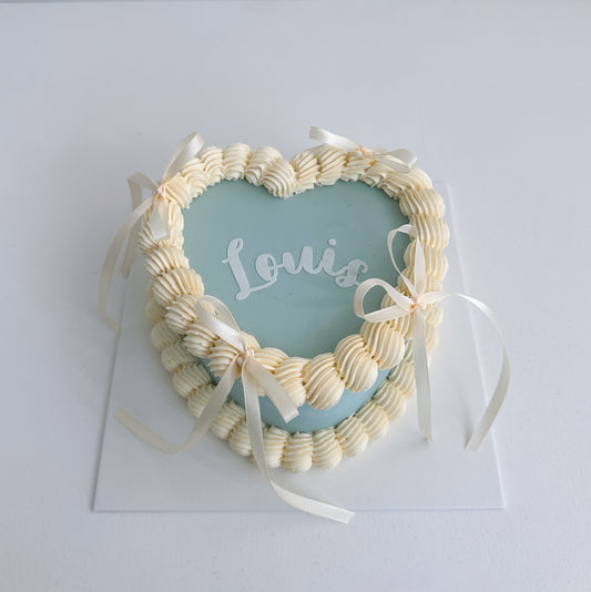 Heart-shaped decorative cake with 'Louis' on a light blue card, surrounded by white ribbons on a light gray background.